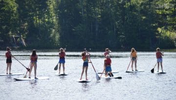 WeHaKee Camp for Girls campers standing on paddle boards on Lake Hunter on a sunny day.