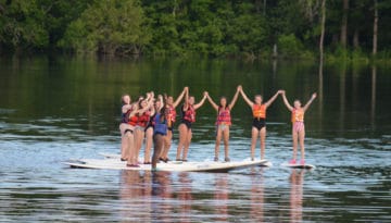 WeHakee Camp for Girls campers holding hands while standing on paddle boards.