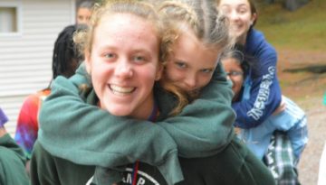 WeHakee Camp for Girls camper giving another camper a piggyback ride while walking amongst other campers.