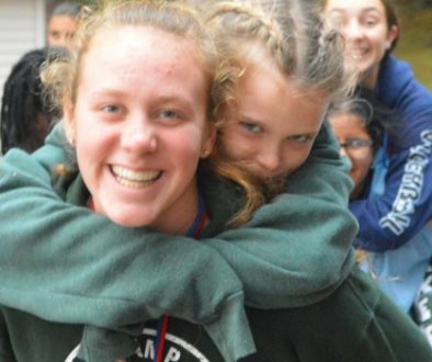 WeHakee Camp for Girls camper giving another camper a piggyback ride while walking amongst other campers.