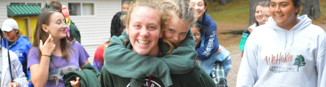 WeHakee Camp for Girls camper giving another camper a piggyback ride while walking amongst other campers. WeHakee Camp for Girls camper giving another camper a piggyback ride while walking amongst other campers.